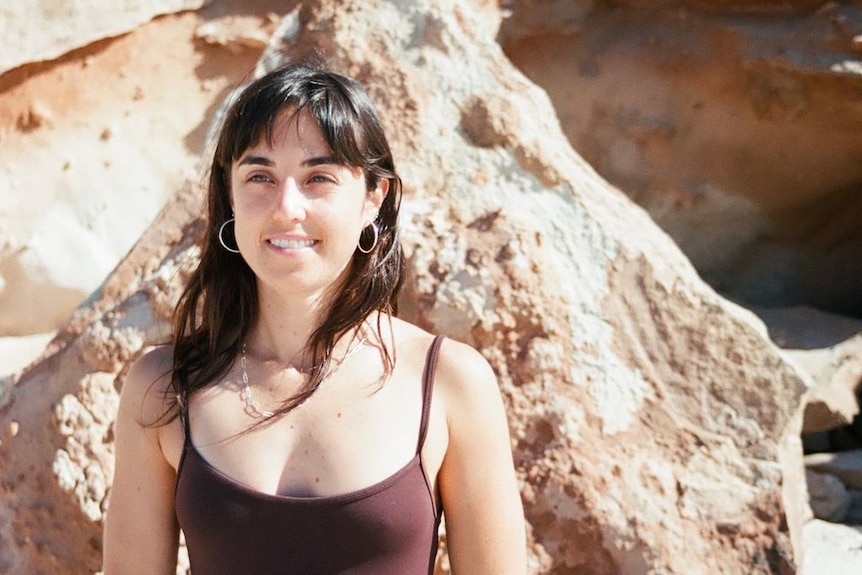 A young woman in brown singlet with long dark brown hair stands in front of a rock on a beach looking off camera.