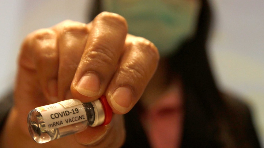 A hand holding a small glass vial containing liquid with a label that says COVID-19 mRNA vaccine