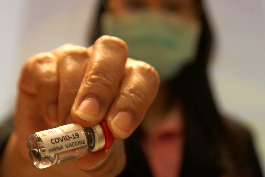 A hand holding a small glass vial containing liquid with a label that says COVID-19 mRNA vaccine