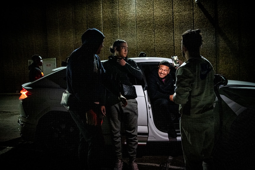 A man sits in a car with the passenger door open at night. Others young men stand around, one taking a photo.
