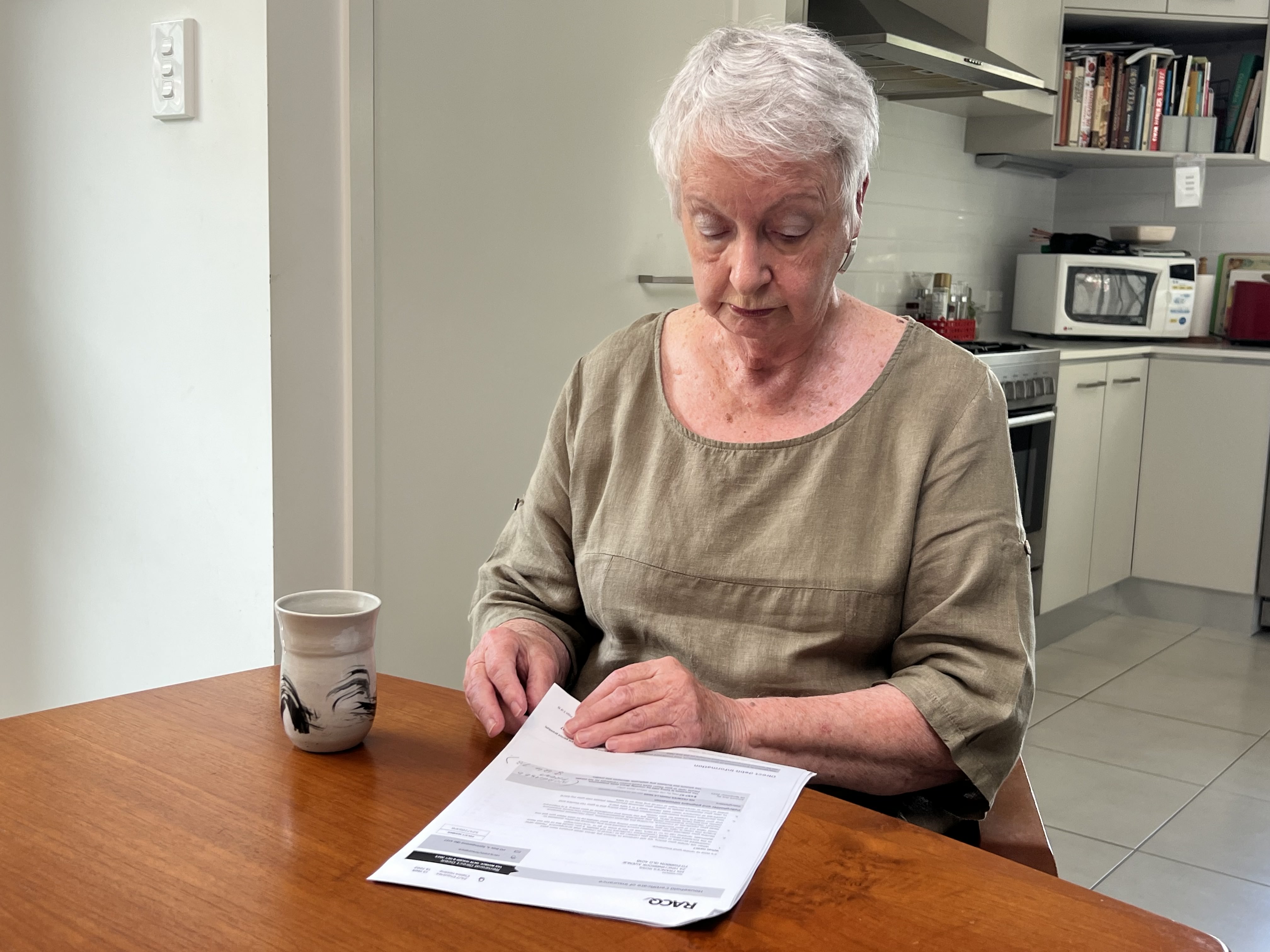 a woman sits at a kitchen table with a cup of tea reading a stack of documents