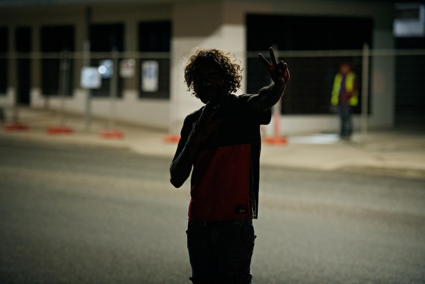 A person in shadows holds a peace sign to the camera. 