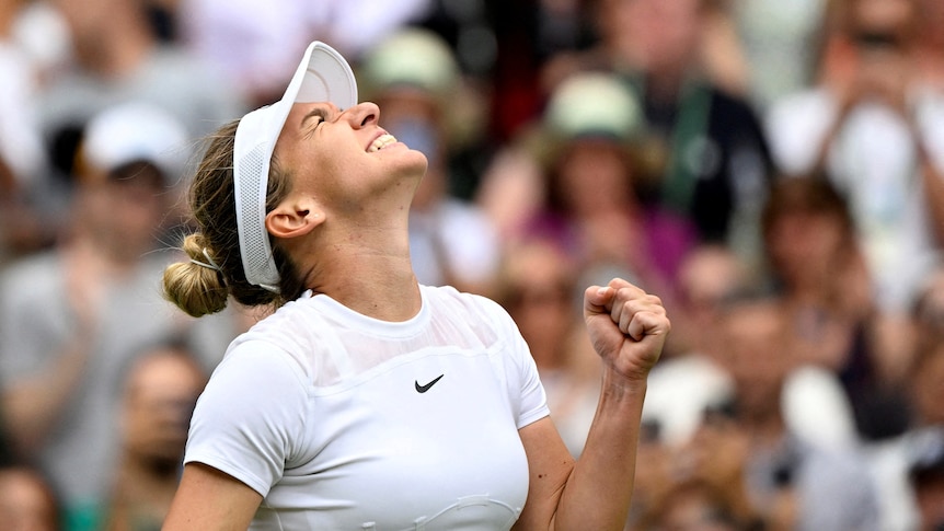 Woman dressed in white tennis clothes smiles and looks up to the sky as she pumps her fist in celebration.