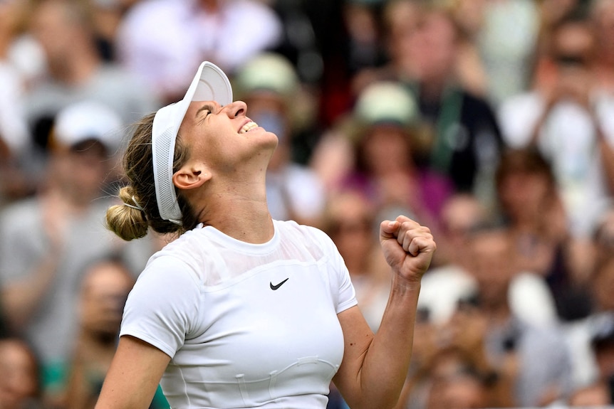Woman dressed in white tennis clothes smiles and looks up to the sky as she pumps her fist in celebration.