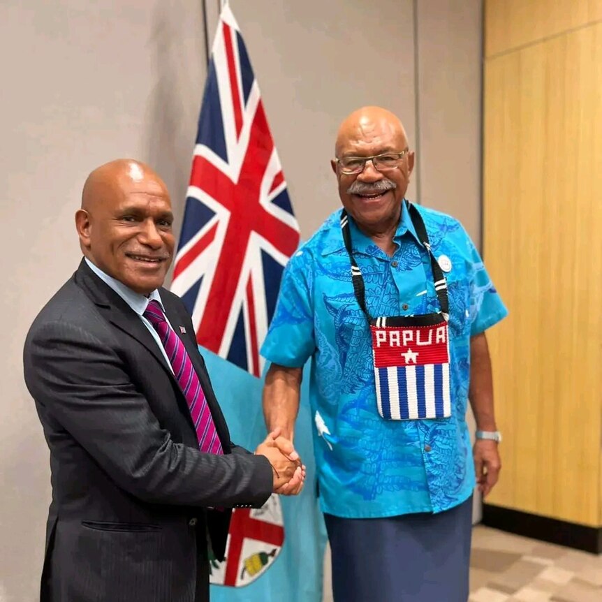 Two men shake hands in front of a Fiji flag.