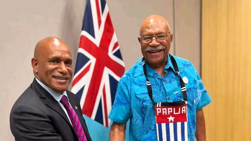 Two men shake hands in front of a Fiji flag.