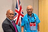 Two men shake hands in front of a Fiji flag.