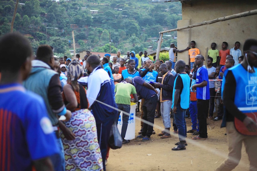 Crowds of Congolese people, many in blue vests, congregate around a plastic tub where people are casting votes.