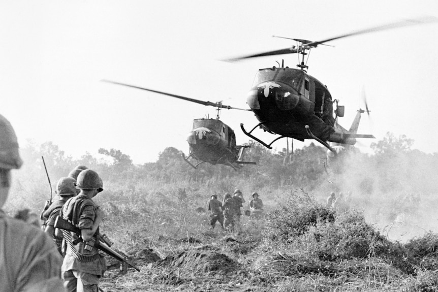 Black and white photo of two helicopters landing in field with soldiers, during Vietnam War.