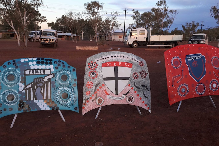 Three car bonnets featuring AFL team logos painted in an Indigenous style, displayed in the WA outback.