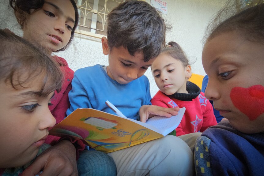 A young boy sits holding a yellow book while young children read over his shoulder.