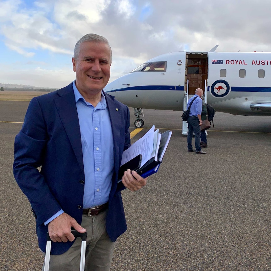 Michael McCormack smiles while wheeling a suitcase towards a plane on the tarmac.