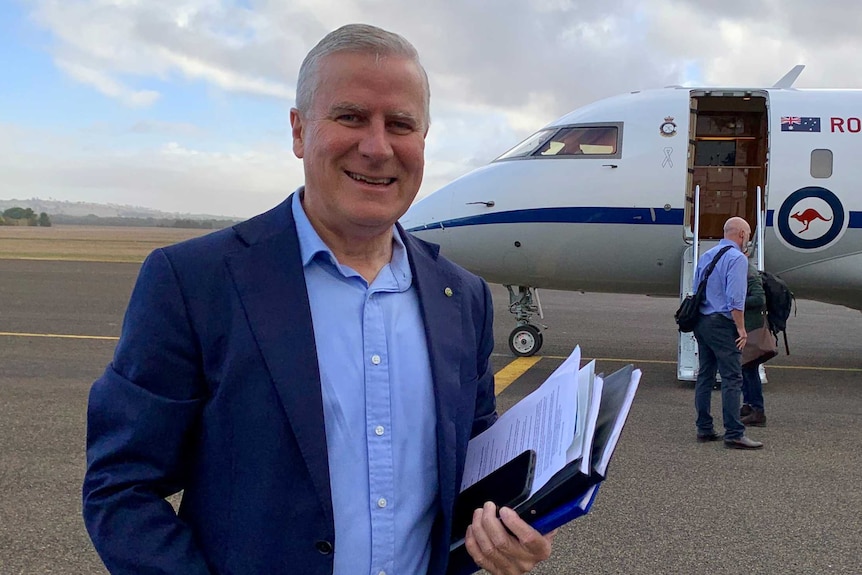 Michael McCormack smiles while wheeling a suitcase towards a plane on the tarmac.
