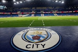 The club emblem of Manchester City is shown in the foreground with its stadium in the background