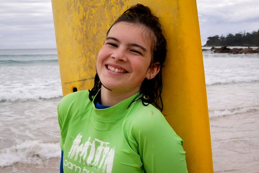 Young girl standing at the edge of beach with surfboard and huge smile