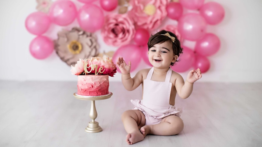 Arianna Maragol in a pink baby overall celebrating her 1st birthday with a pink iced cake and pink balloons and flowers