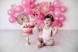 Arianna Maragol in a pink baby overall celebrating her 1st birthday with a pink iced cake and pink balloons and flowers