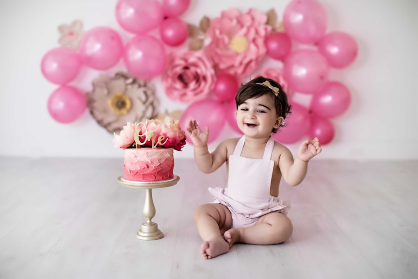 Arianna Maragol in a pink baby overall celebrating her 1st birthday with a pink iced cake and pink balloons and flowers