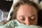 A woman with blond hair and her eyes closed lies on a bed with a cat behind her.