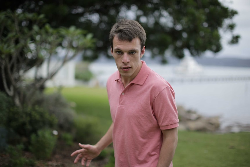 A young man in a peach-coloured tshirt looking at the camera with a beach in the background