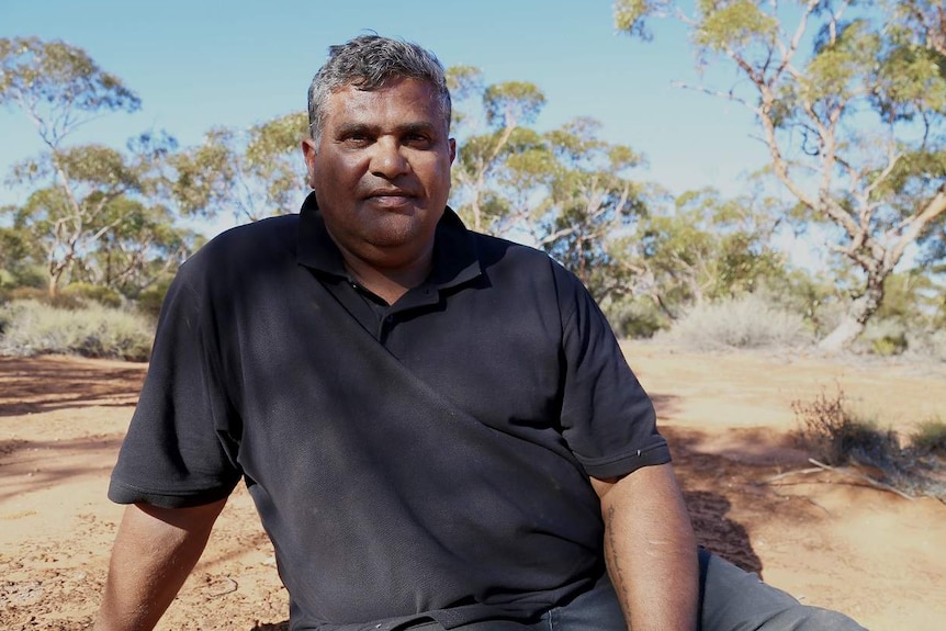 An Anangu man sits on land with trees in the background.