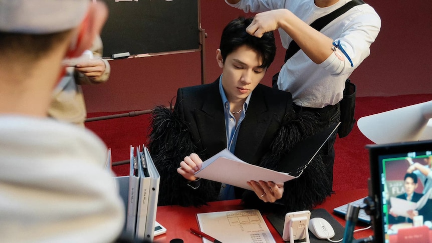 A young man reads paper while two other people help him with his makeup. 