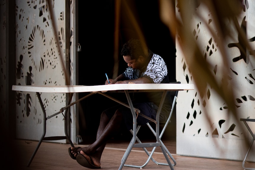 A man wearing a T-shirt and shorts sitting and drawing at a trestle table outside a building. 