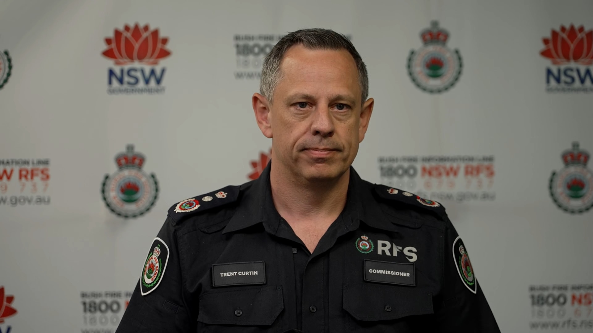 Trent wears a dark uniform and has short dark hair and a serious expression.