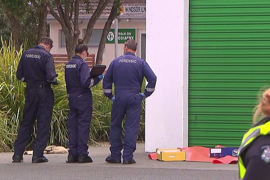 Three police in navy jumpsuits with the word "forensic" on the back examine a crime scene.