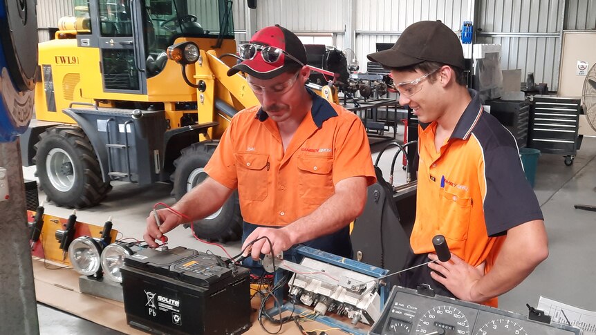 man showing younger man how to connect devices to a battery, in workshop with Forklift in the background