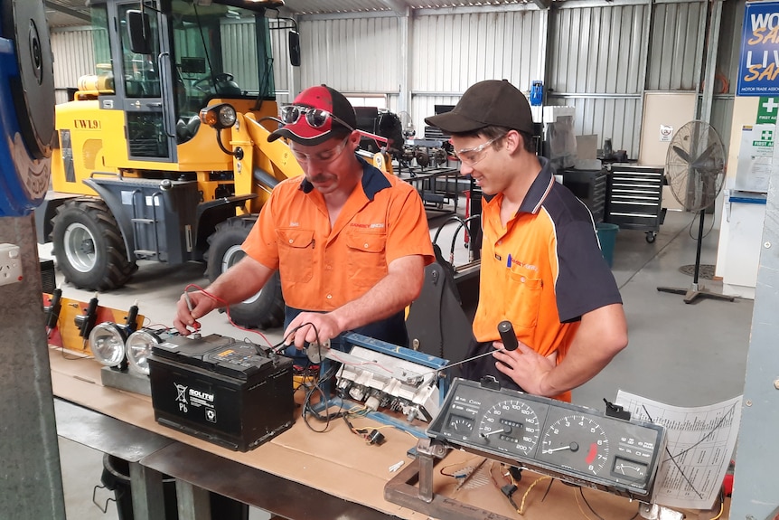 man showing younger man how to connect devices to a battery, in workshop with Forklift in the background