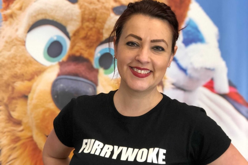 A woman in a black shirt with the word 'furrywoke' on it smiles and stands in front of a lion costume