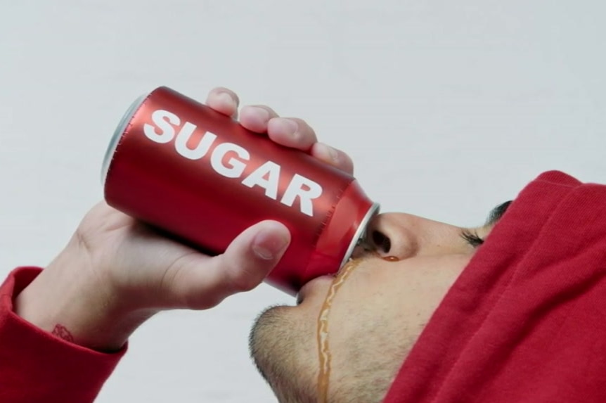 A man in a red hoodie drinks from a red can labelled 'sugar', with a cola-coloured liquid running down his cheek.