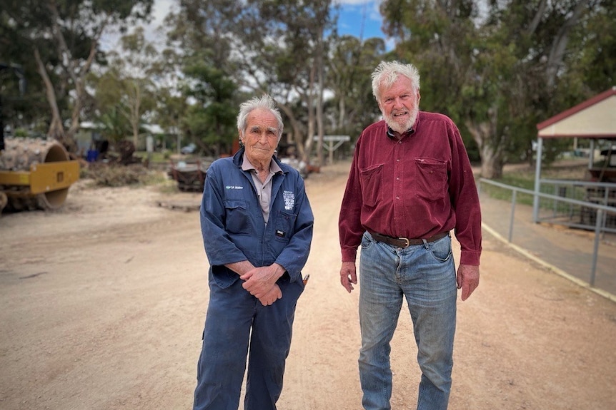 Two men, wearing a blue jumpsuit and a red shirt, stand outside on a dirt road.