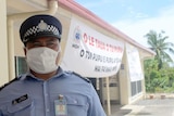 A Samoan police officer stands wearing a white medical mask in front of a building with a white banner.