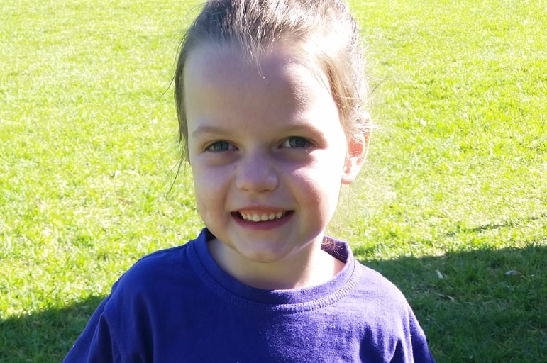 Ella stands on a lawn smiling. She is wearing a purple t-shirt and tutu.