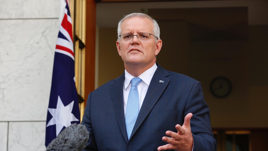 Scott Morrison in a suit and light blue tie in front of a microphone with an Australian flag behind him