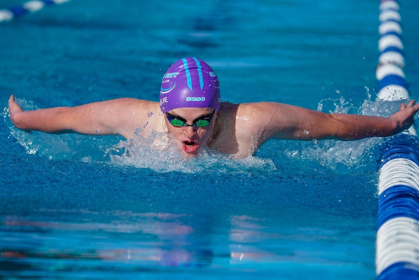 A teenaged boy wearing a purple and blue swim cap and goggles swimming in a pool lane.