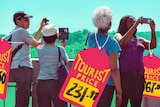 A photo of ageing tourists taking photos has been overlaid with large hand-drawn price tags marked with 'tourist prices'