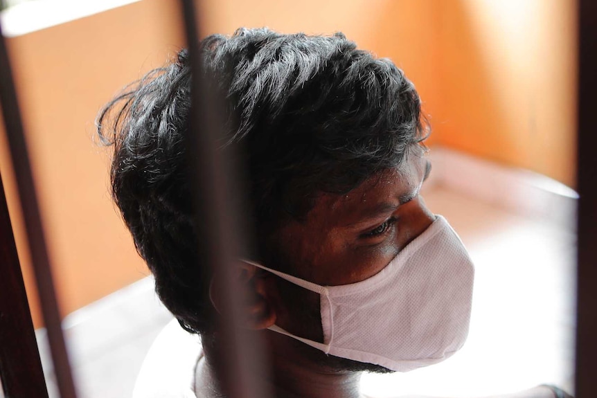 Dark haired man is seen through window wearing a white mask