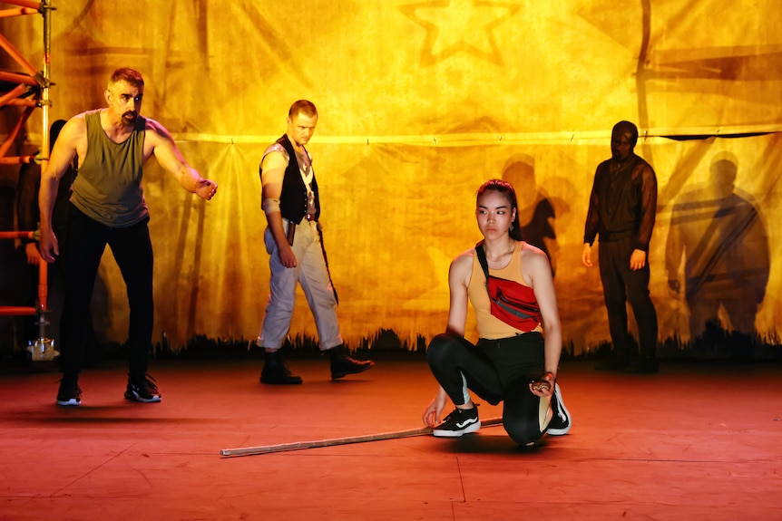 An Asian woman crouches on a stage, a bang at her feet. Behind her stands an Arab man. A white man skulks behind them both.