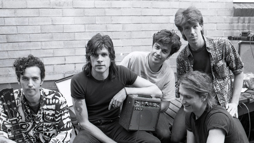 black and white photo of five smiling men sitting outdoors with a very small guitar amp