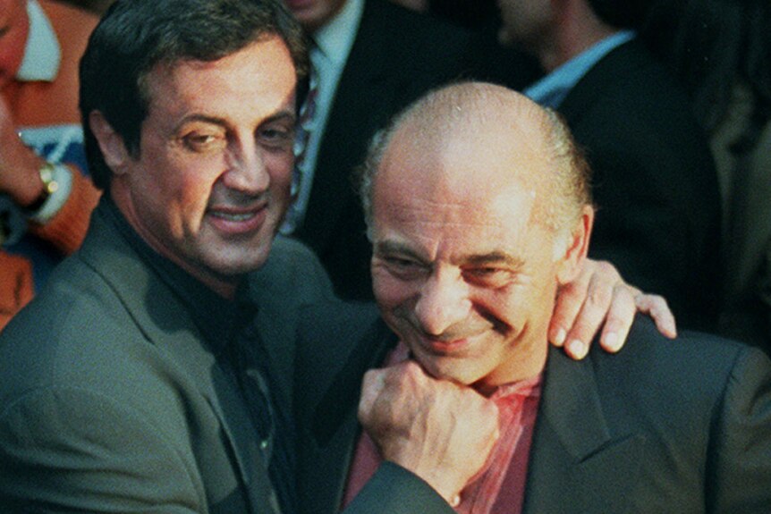 Sylvester Stallone jokingly holds his fist up to Burt Young's face. Both of them wear suits.