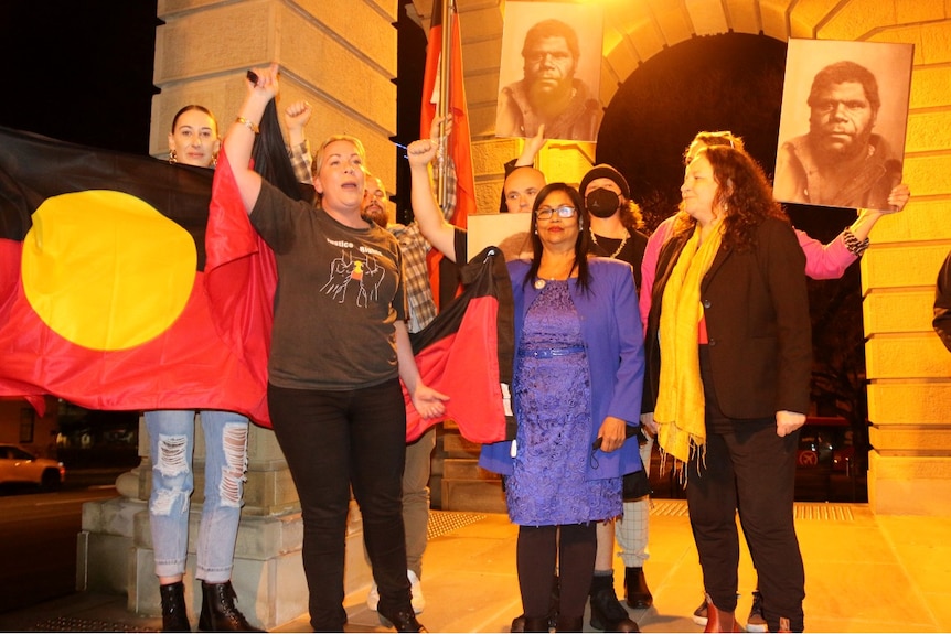 A group of Indigenous people hold an Aboriginal flag and cheer.