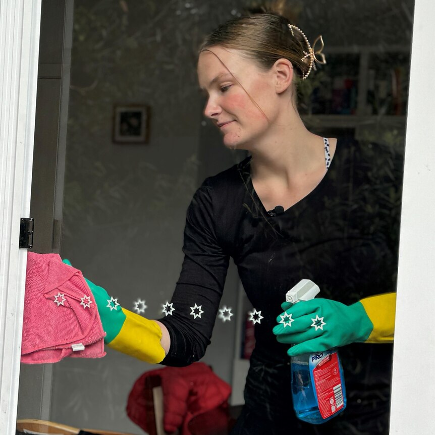 A young woman wearing a black shirt and green and yellow gloves cleans a glass sliding door from inside a house.