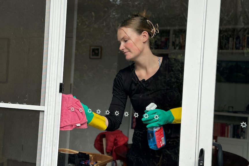 A young woman wearing a black shirt and green and yellow gloves cleans a glass sliding door from inside a house.