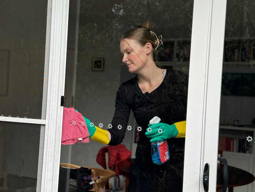 A young woman wearing a black shirt and green and yellow gloves cleans a glass sliding door from inside a house.