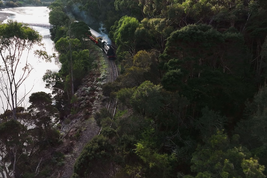 A steam train making its way around a bend with forest on one side and a river on the other.