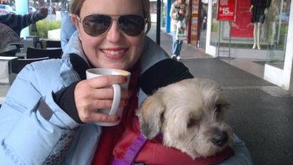 A woman wearing sunglasses sits outside a cafe with a coffee, and a small dog on her lap.