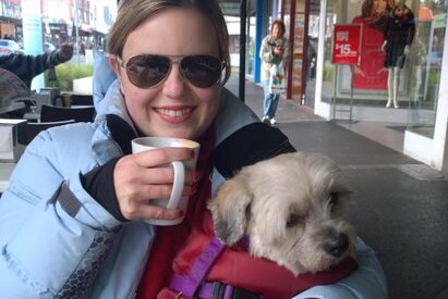 A woman wearing sunglasses sits outside a cafe with a coffee, and a small dog on her lap.